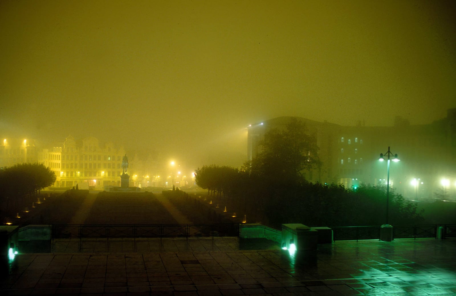 Une esplanade en pente bordée de bancs et de lumières vertes et jaunes, la ville se devinant dans la brume