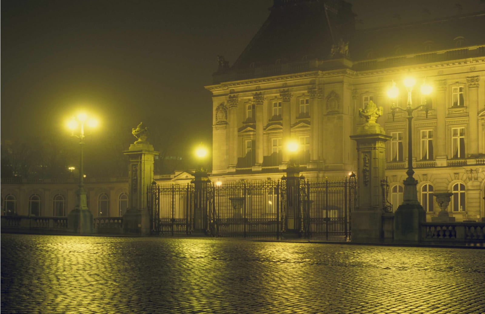 Une grille monumentale devant un palais éclairé, au bord d’une place mouillée