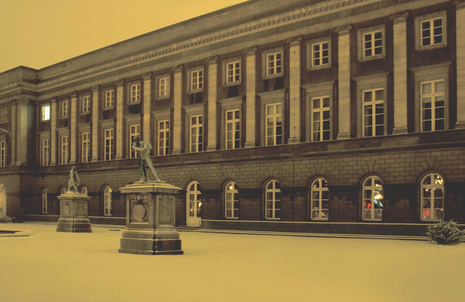 La longue façade d’un palais devant une esplanade enneigée, des statues au premier plan