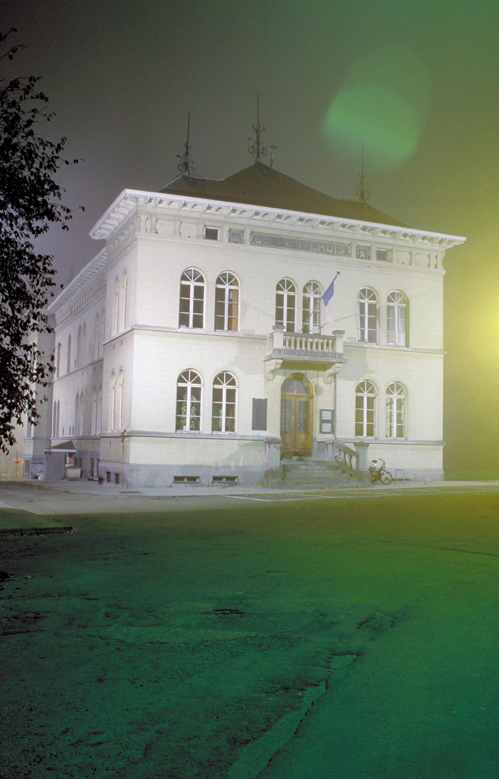 Un édifice blanc à fronton éclairé dans la nuit, cerné d’ombres vertes