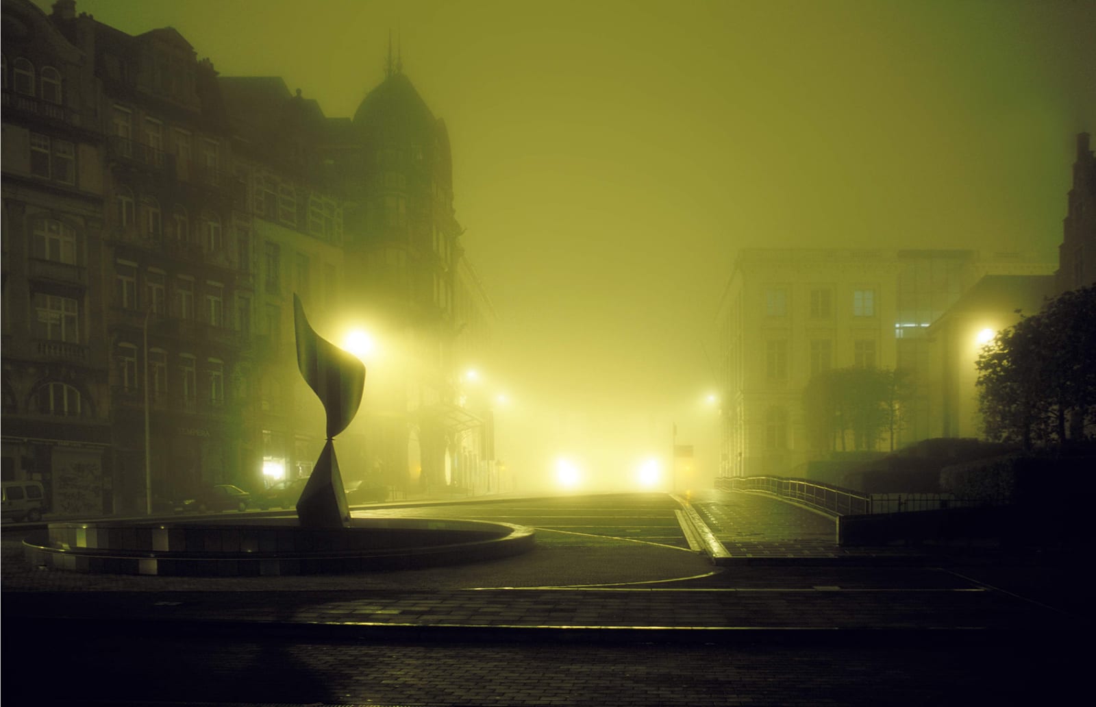 Une sculpture abstraite au centre d’un rond-point désert, dans la brume verte