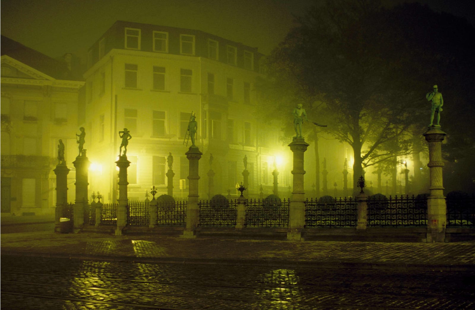 Des statues juchées sur des colonnes le long d’une grille, devant des façades noyées de brume